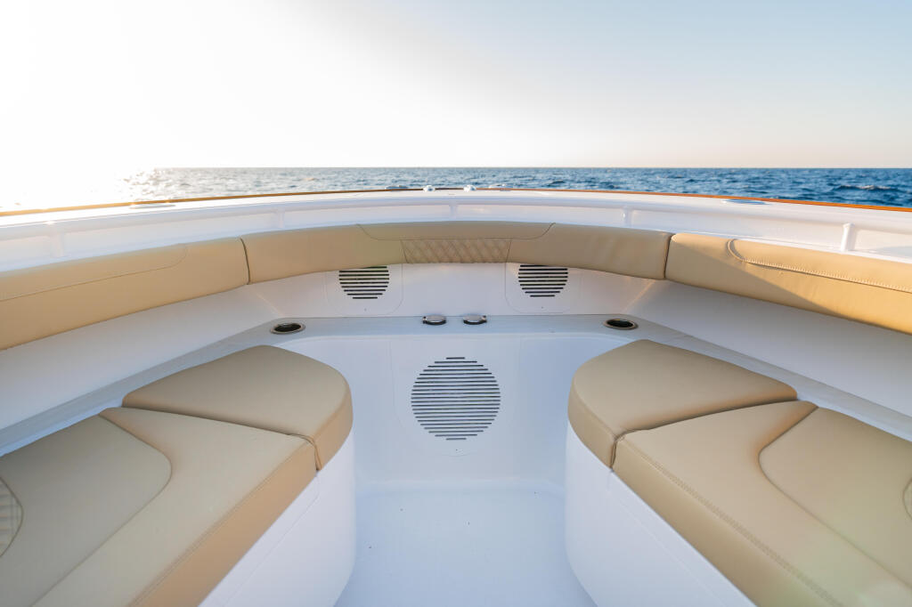 Curved bow seating with tan cushions and audio speakers visible on white fiberglass deck overlooking calm blue water