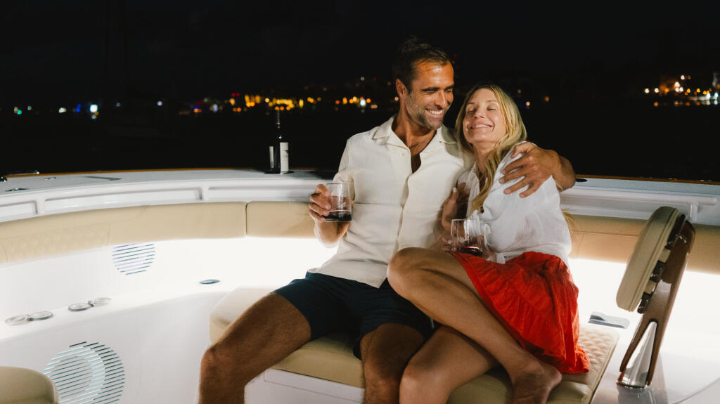 Couple enjoying drinks together on a boat's helm at night with marina lights in the background