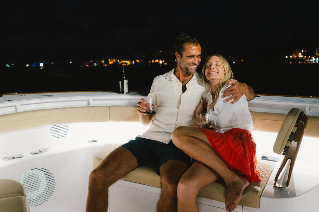 Couple enjoying drinks together on a boat's helm at night with marina lights in the background